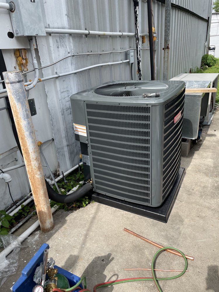 A gray HVAC condenser unit sits on a concrete pad next to a corrugated metal wall with tools nearby.