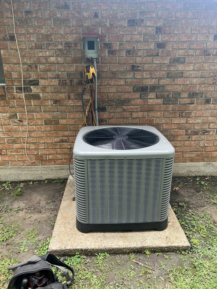 An outdoor air conditioning unit sits on a concrete slab against a brick wall with an electrical disconnect box above.