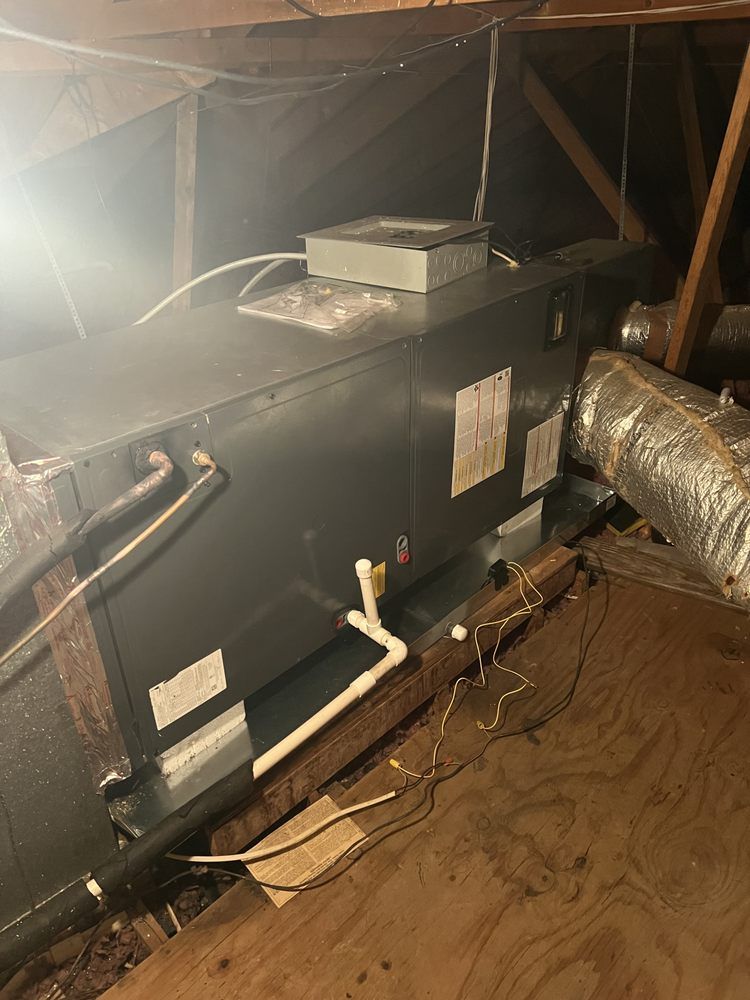 An HVAC air handler unit installed in an attic, featuring metal casing, attached ducts, and white PVC drain piping.