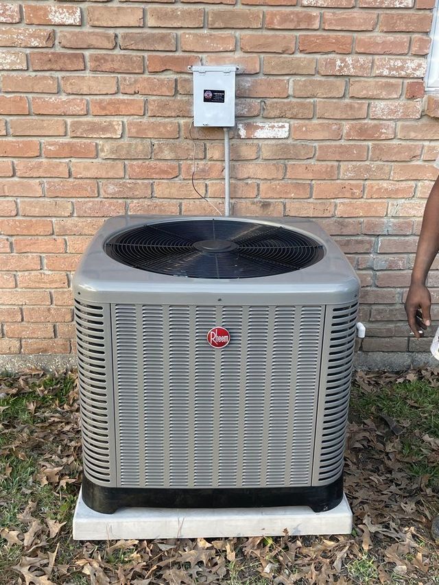 A grey Rheem HVAC outdoor unit sits on a concrete pad against a brick wall, with a disconnect box mounted above it.