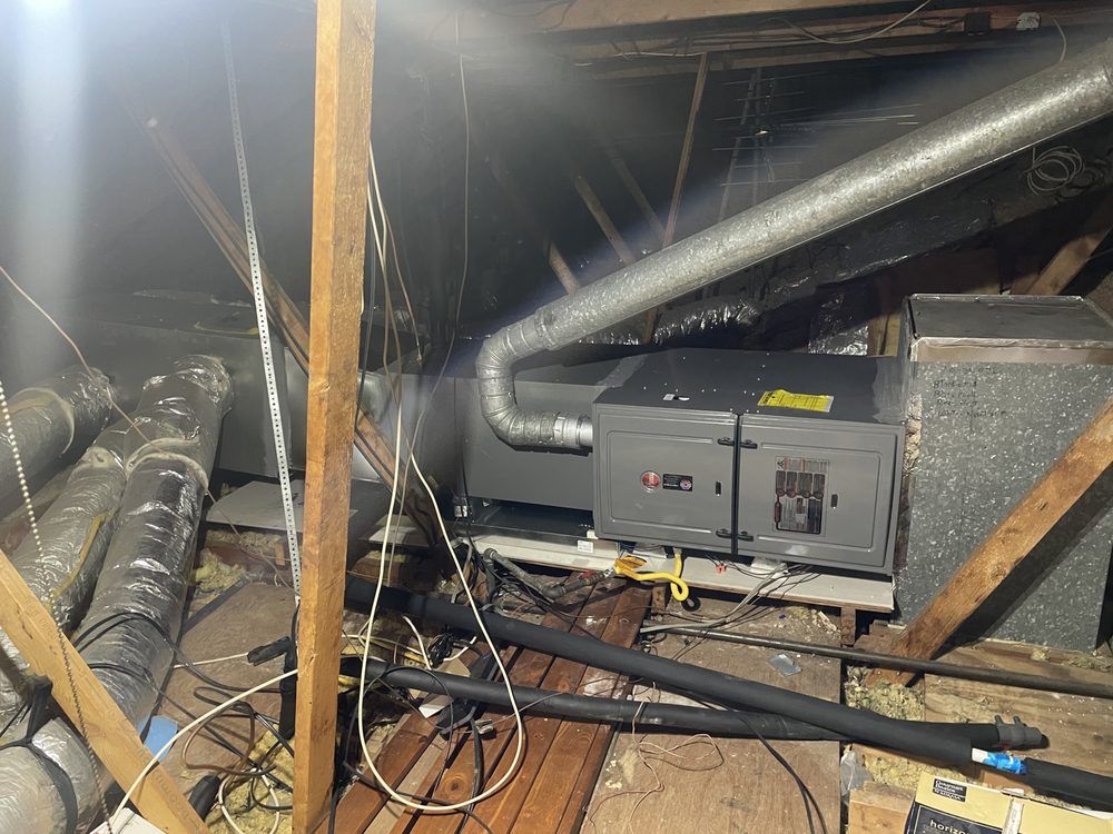 A grey HVAC air handler unit connected to silver metal ductwork inside a wooden attic space.