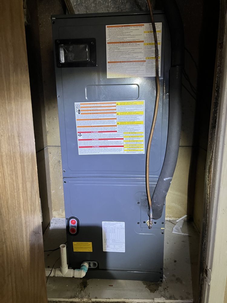 A gray HVAC air handler unit standing in a small closet, with a white PVC drain pipe connected to its base.