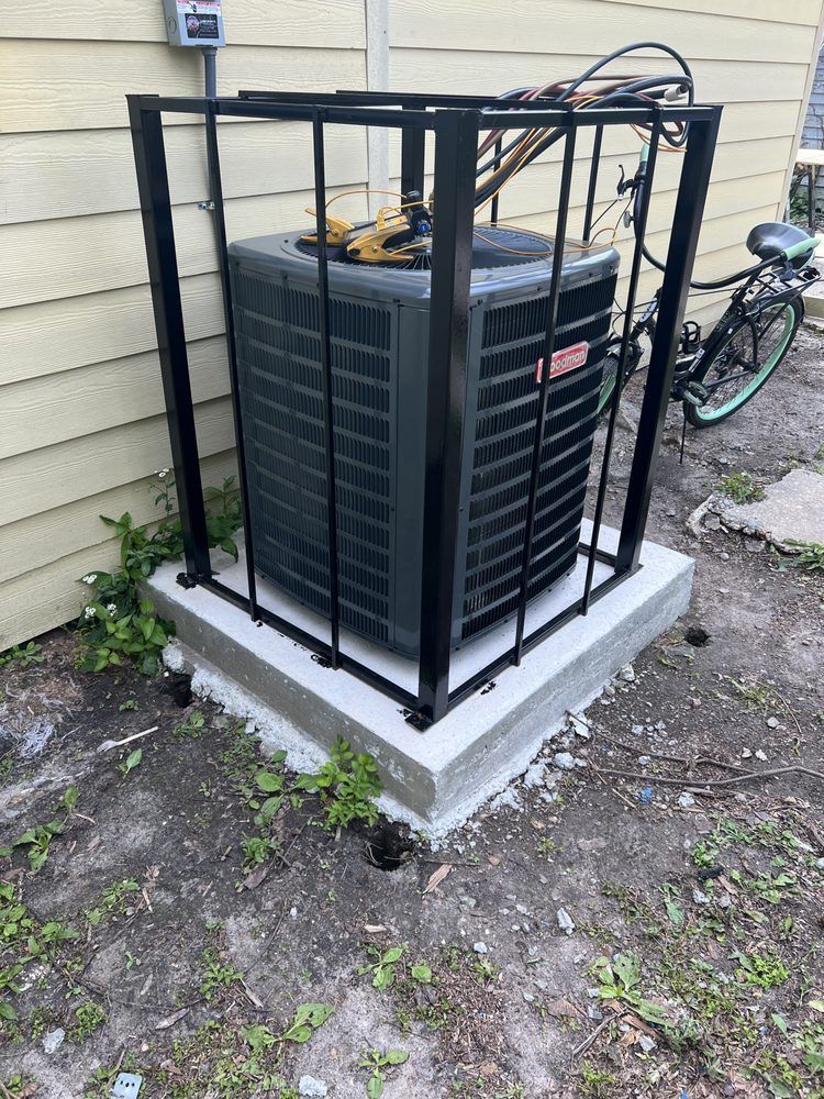 An outdoor AC unit enclosed in a black metal security cage, sitting on a concrete pad next to a house wall and a bicycle.
