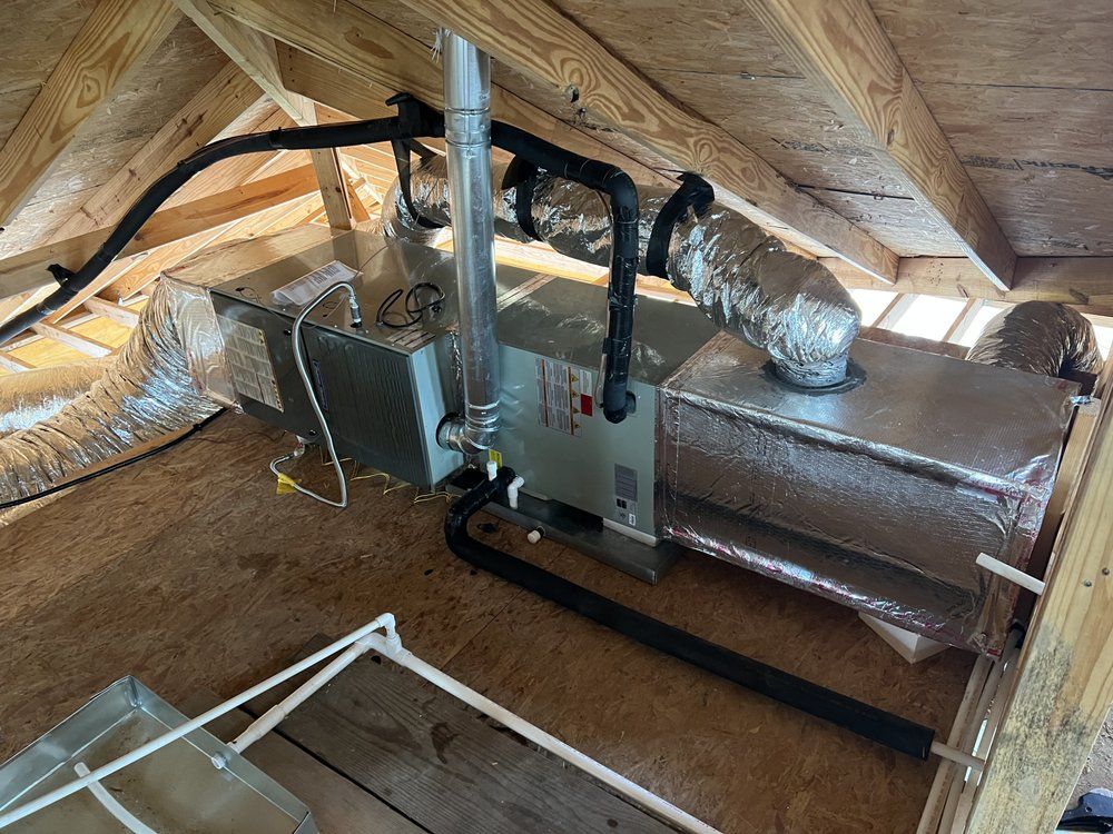 A horizontal HVAC air handler unit with ductwork and plumbing installed in an unfinished wooden attic.