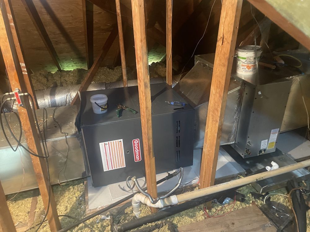 A dark gray HVAC furnace unit installed in an attic space, connected to metal ductwork and resting on a white platform.