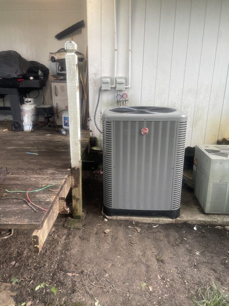 A grey Rheem outdoor HVAC unit sits on a concrete pad next to a wooden deck and a white vertical-siding wall.