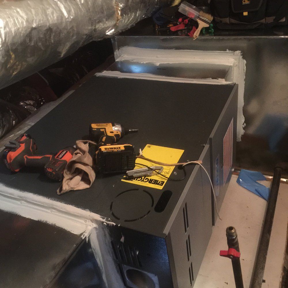 A dark gray HVAC air handler unit sits in an attic space, with tools and a yellow label on top and sealed ductwork nearby.