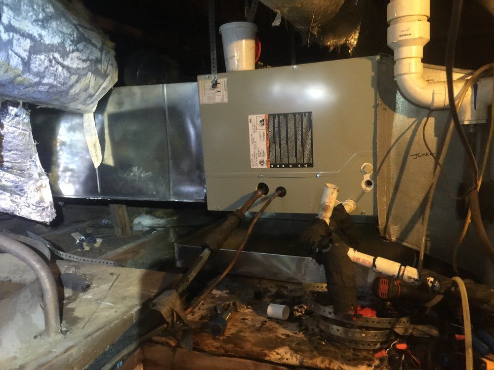 A grey HVAC air handler unit installed in a dark attic space, connected to metal ductwork and white PVC piping.