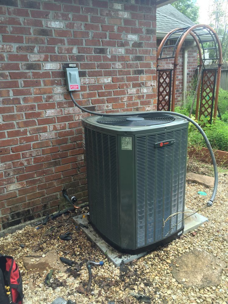 A dark gray HVAC unit sits on a concrete pad against a brick wall, connected to a wall-mounted electrical disconnect box.