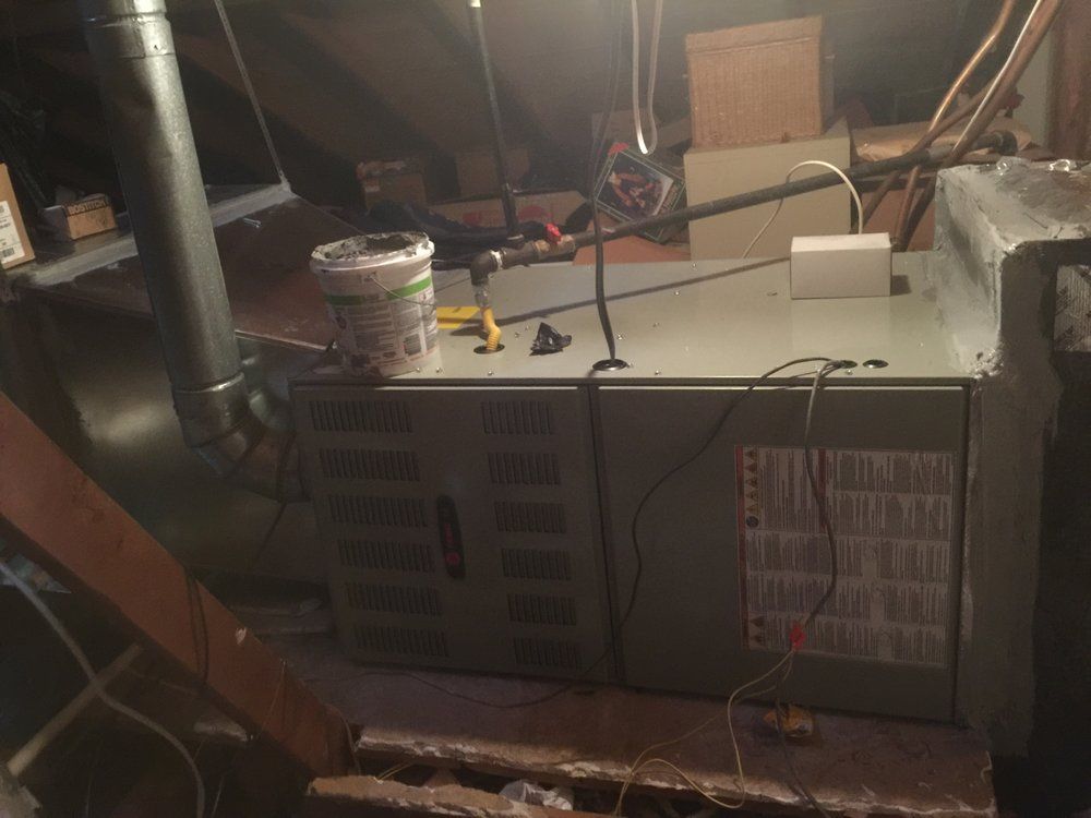 A gray HVAC unit located in a dimly lit attic, with a metal exhaust pipe and gas lines connected to the top.