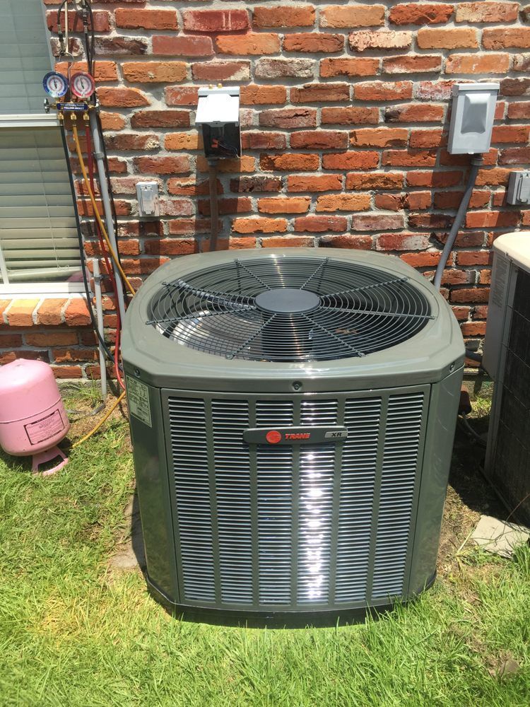 A Trane air conditioning unit sits on grass next to a brick wall with attached gauges and electrical boxes.