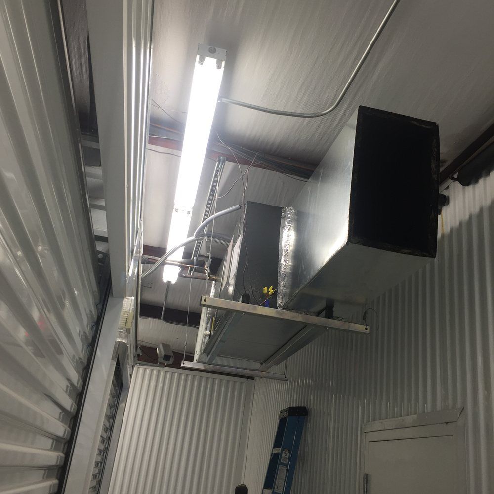 An industrial ceiling-mounted HVAC unit with metal ducting, suspended in a room with corrugated metal walls.