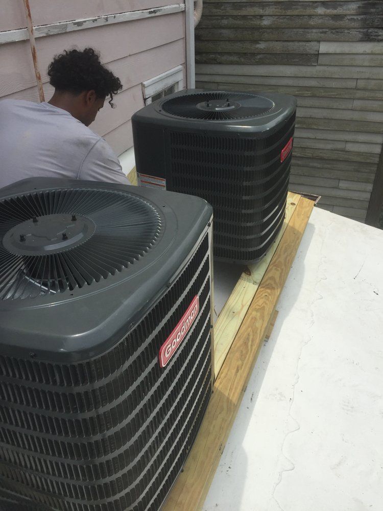 A technician works on two Coleman air conditioning units mounted on a wooden platform on a flat rooftop.
