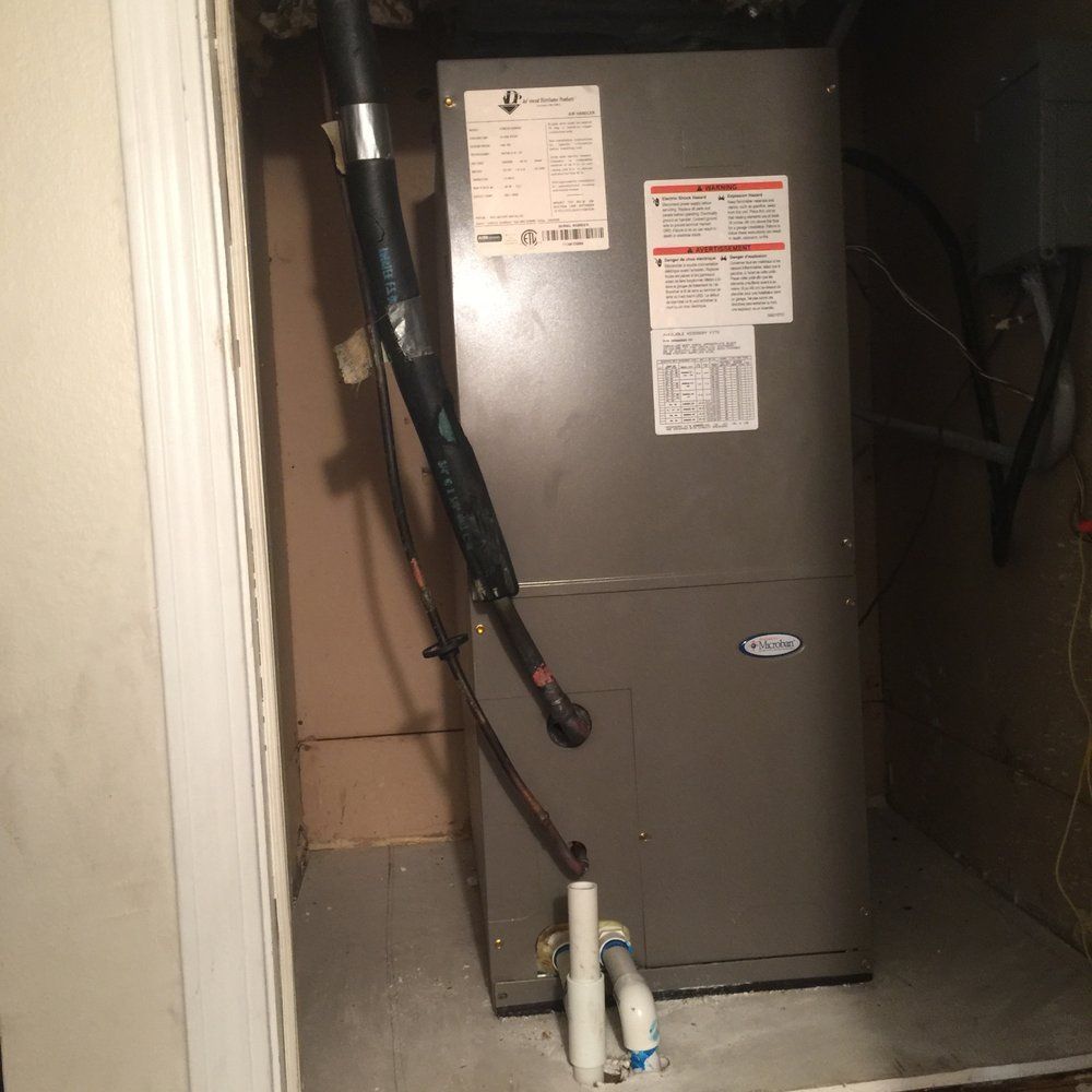 A gray HVAC unit in a utility closet with pipes and electrical wiring attached.