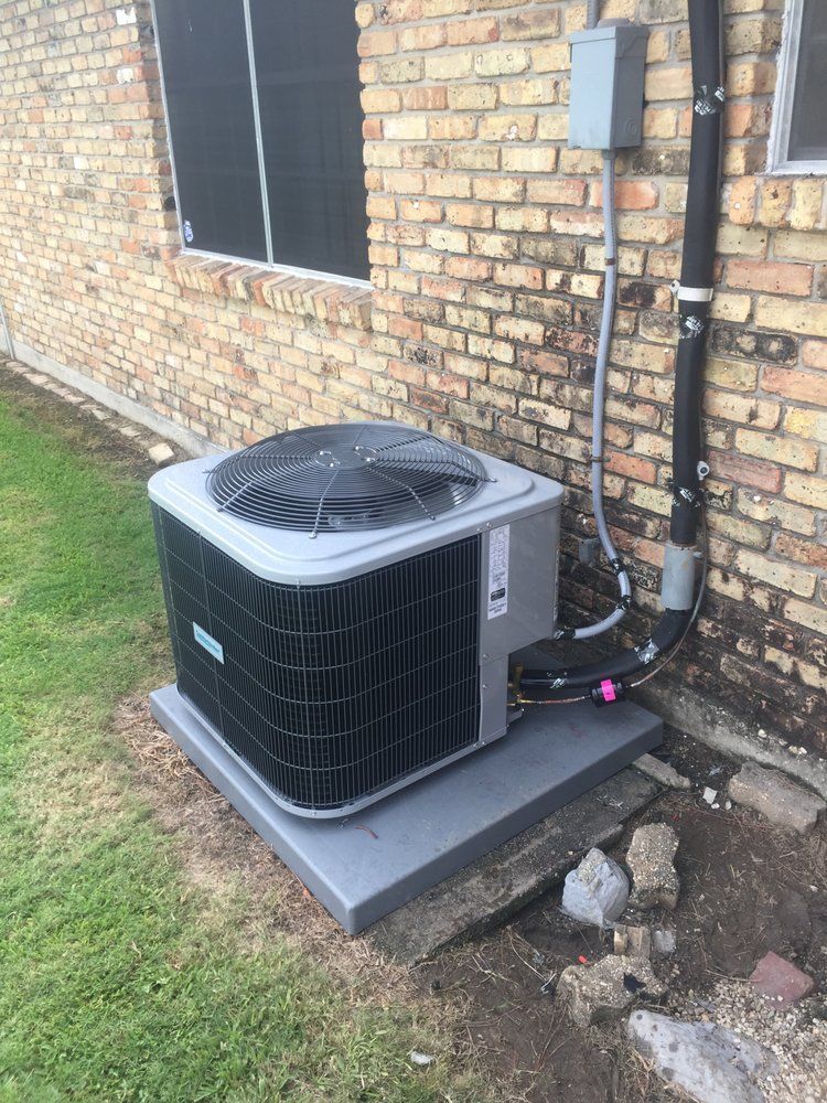 A grey outdoor air conditioning unit sits on a concrete pad against a brick wall next to a window and utility conduit.