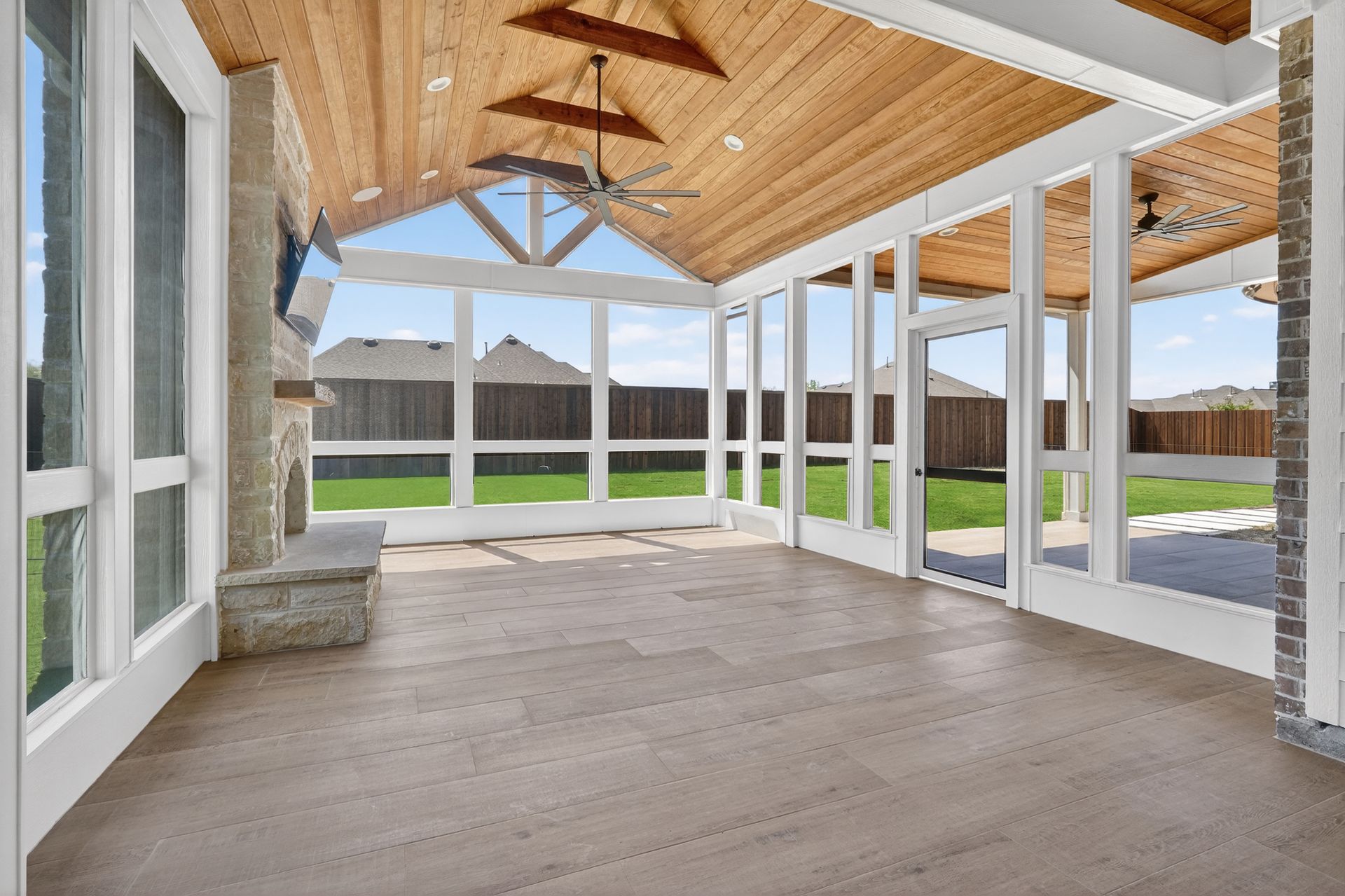An airy, covered patio with vaulted wood-plank ceilings, a stone fireplace, and large glass windows overlooking a yard.