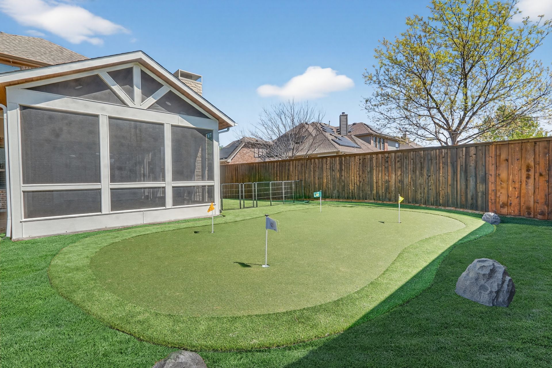 A residential backyard with a synthetic putting green, a screened-in porch, a wooden fence, and decorative rocks.