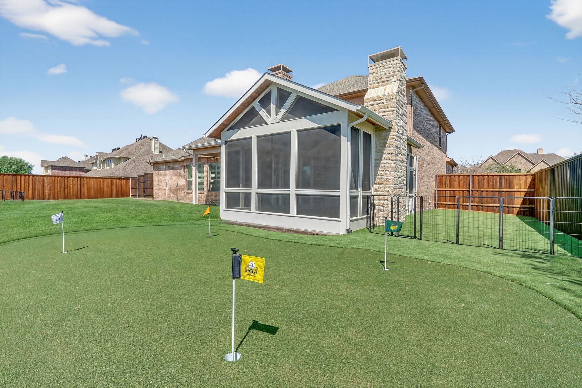 Backyard putting green with yellow and blue flags, featuring a stone house with a screened-in porch under a blue sky.