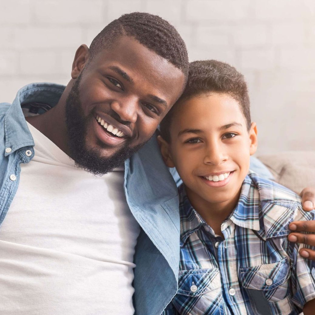 Father and son smiling, arms around each other. The man has a beard. They are indoors.