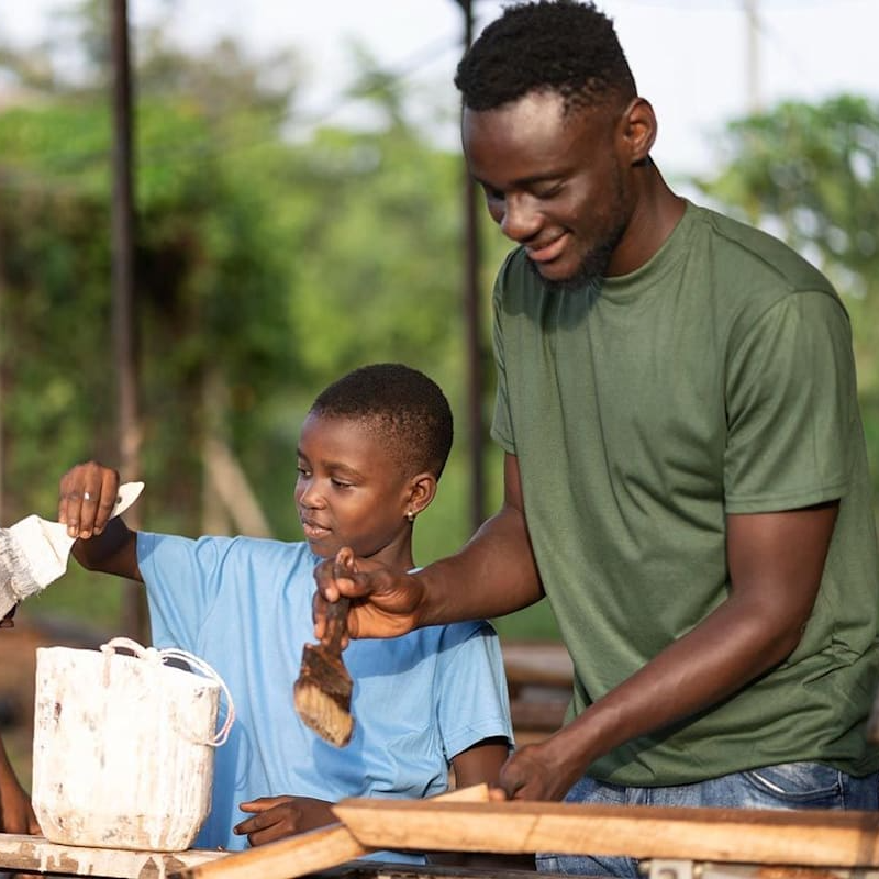 Man and child painting wood together outdoors, smiling.