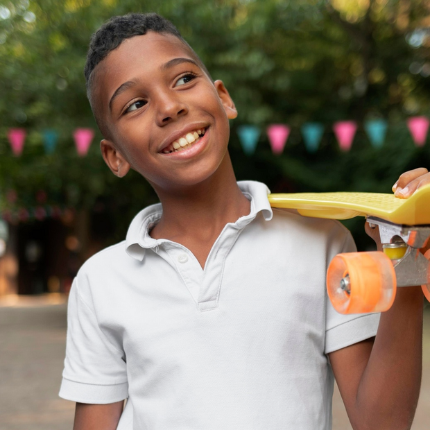 Young Black boy smiles while holding a yellow skateboard. Outdoor setting with colorful bunting.