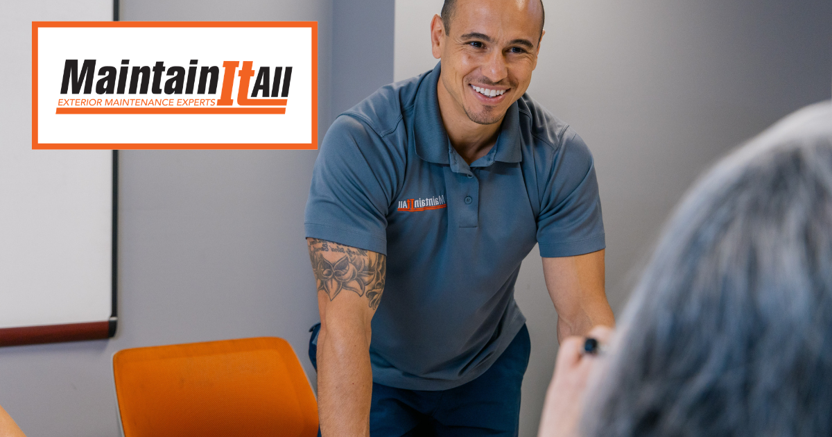 A smiling person wearing a grey polo shirt with a MaintainItAll logo leans over an orange chair in an office setting.