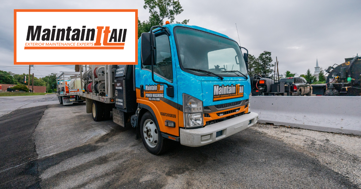 A blue and orange MaintainItAll work truck parked on a gravel lot next to concrete barriers.