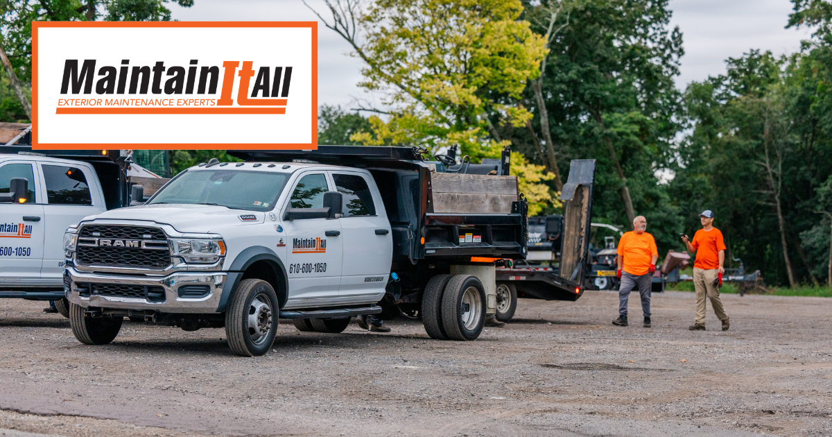 Two people in orange work shirts stand near white dump trucks with the MaintainItAll logo on a gravel lot.