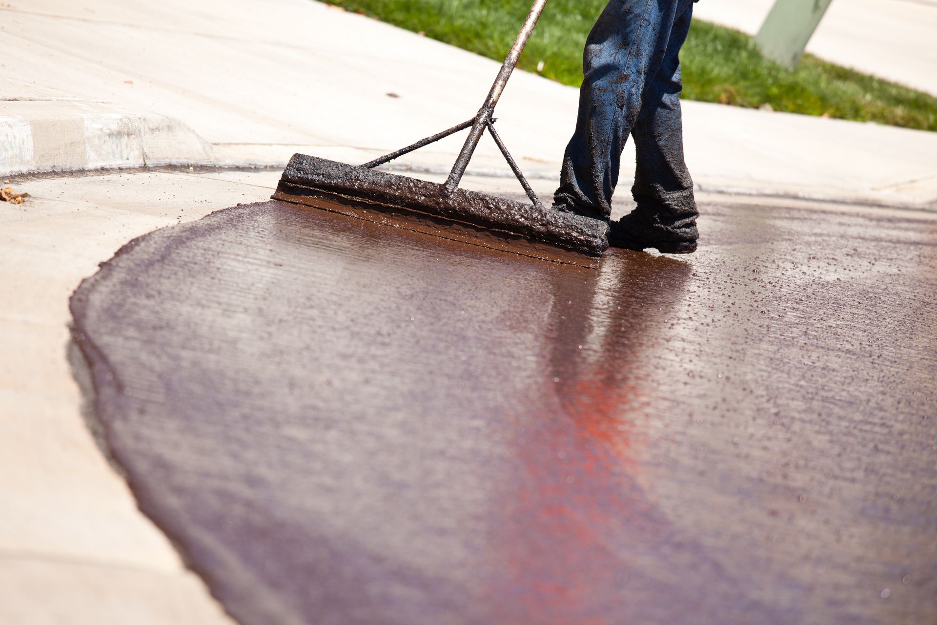 A person uses a long-handled squeegee to spread black asphalt sealer over a residential driveway.