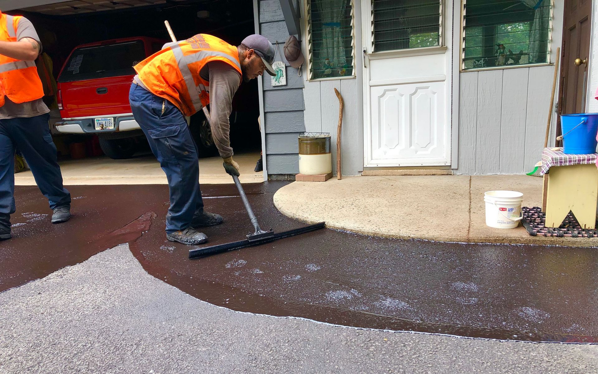 Two workers in orange safety vests use a squeegee to apply black sealant to a driveway in front of a house and garage.