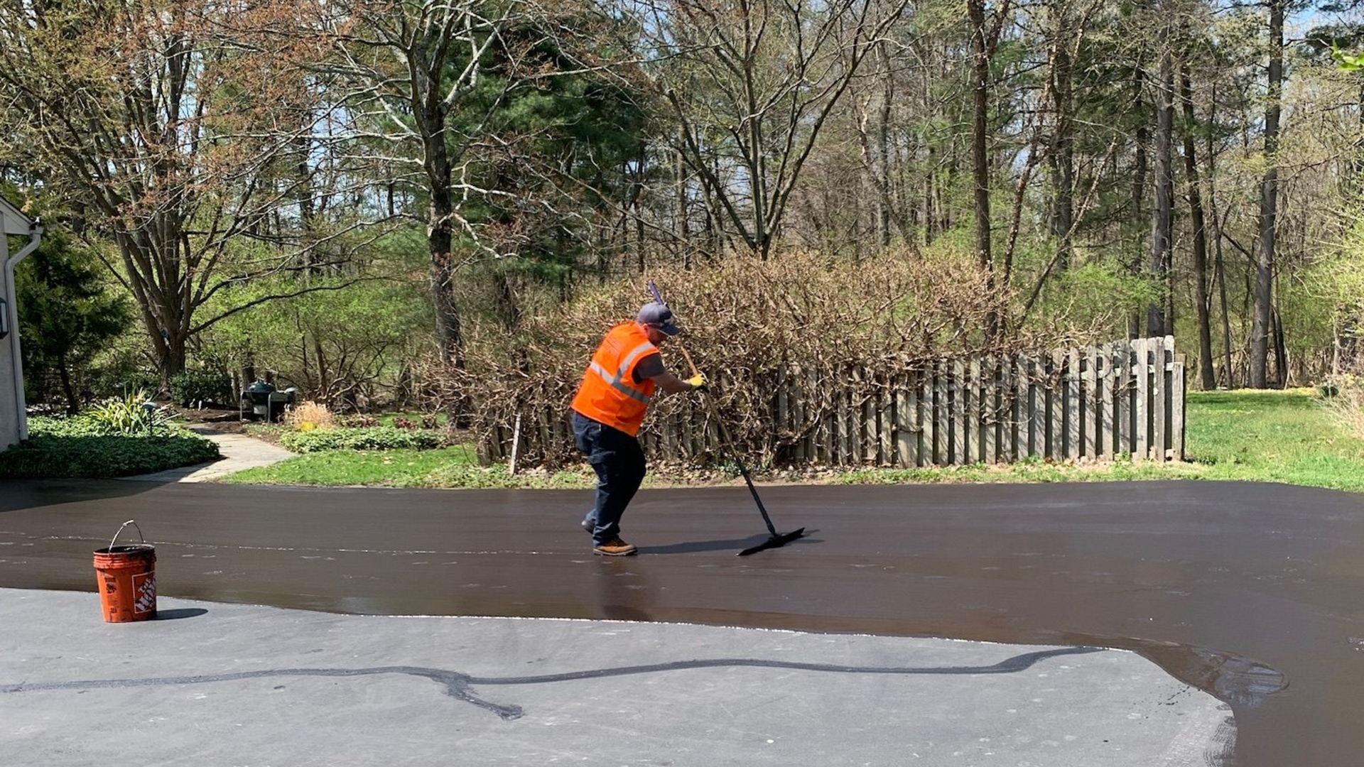 A person in a high-visibility orange vest uses a squeegee to apply black asphalt sealer to a residential driveway.
