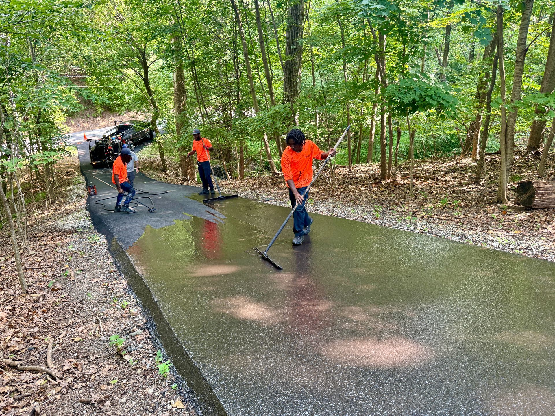 Three workers in orange high-visibility shirts clear water from a paved driveway in a wooded area using squeegees.