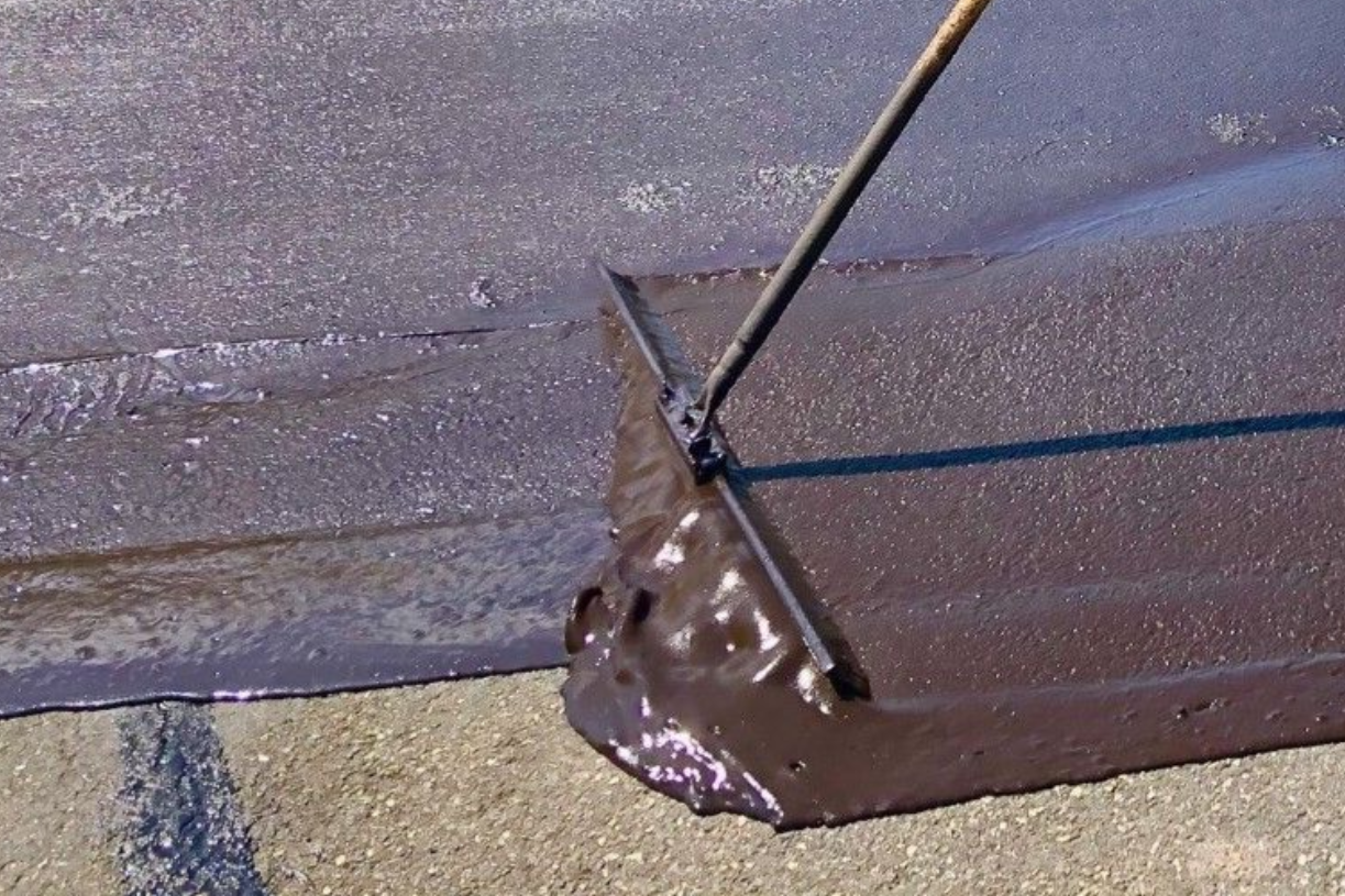 Applying a thick, dark driveway sealant with a squeegee on an asphalt surface.