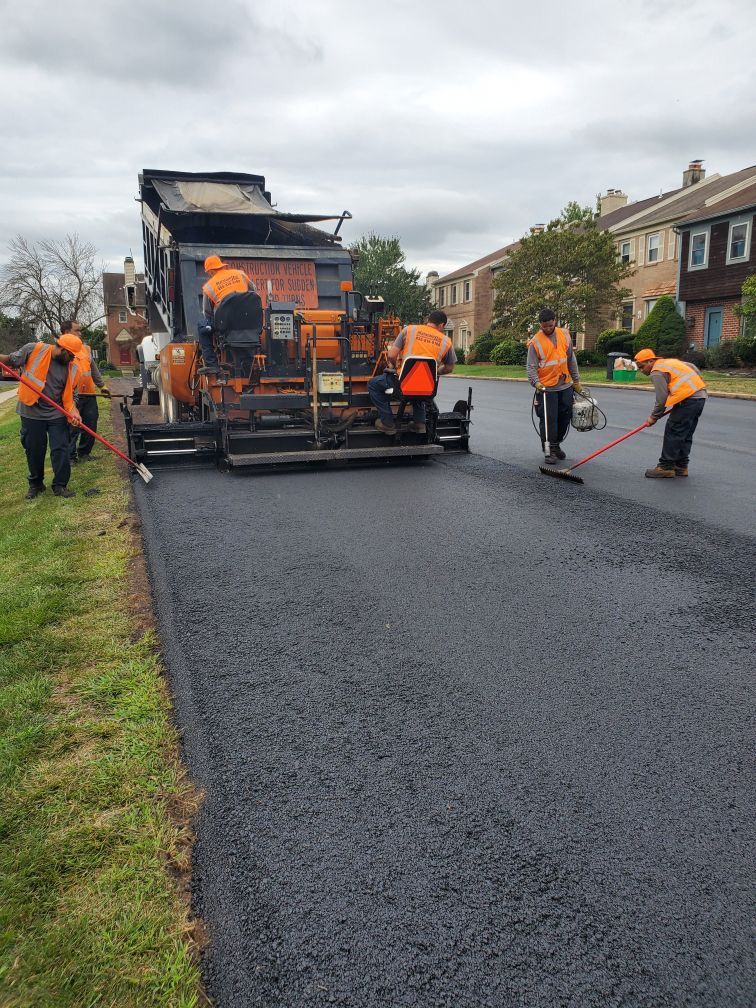 Construction crew in orange safety gear using a paving machine to lay fresh asphalt on a residential street.