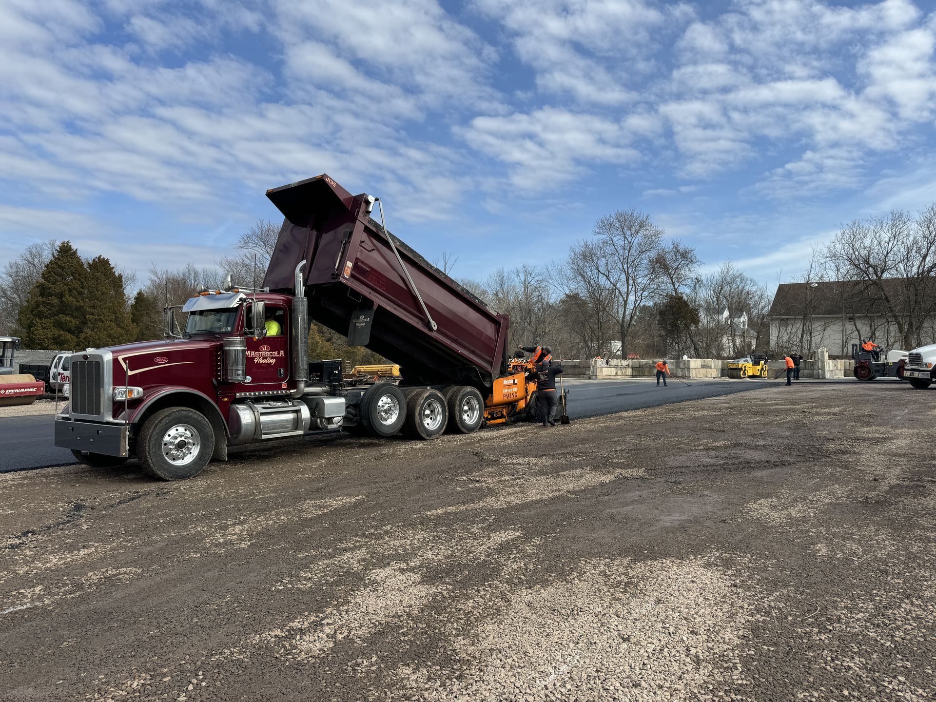 A maroon dump truck raises its bed to feed asphalt into a paving machine on a construction site under a blue, cloudy sky.