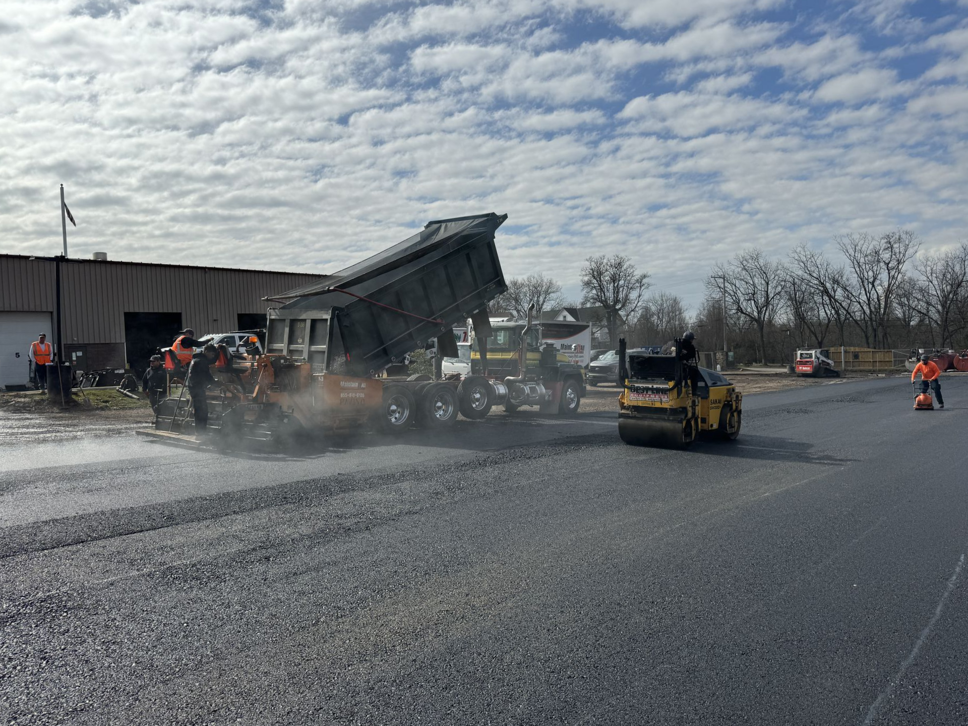 A dump truck with its bed raised and a yellow road roller paving a gravel lot near a building under a cloudy sky.