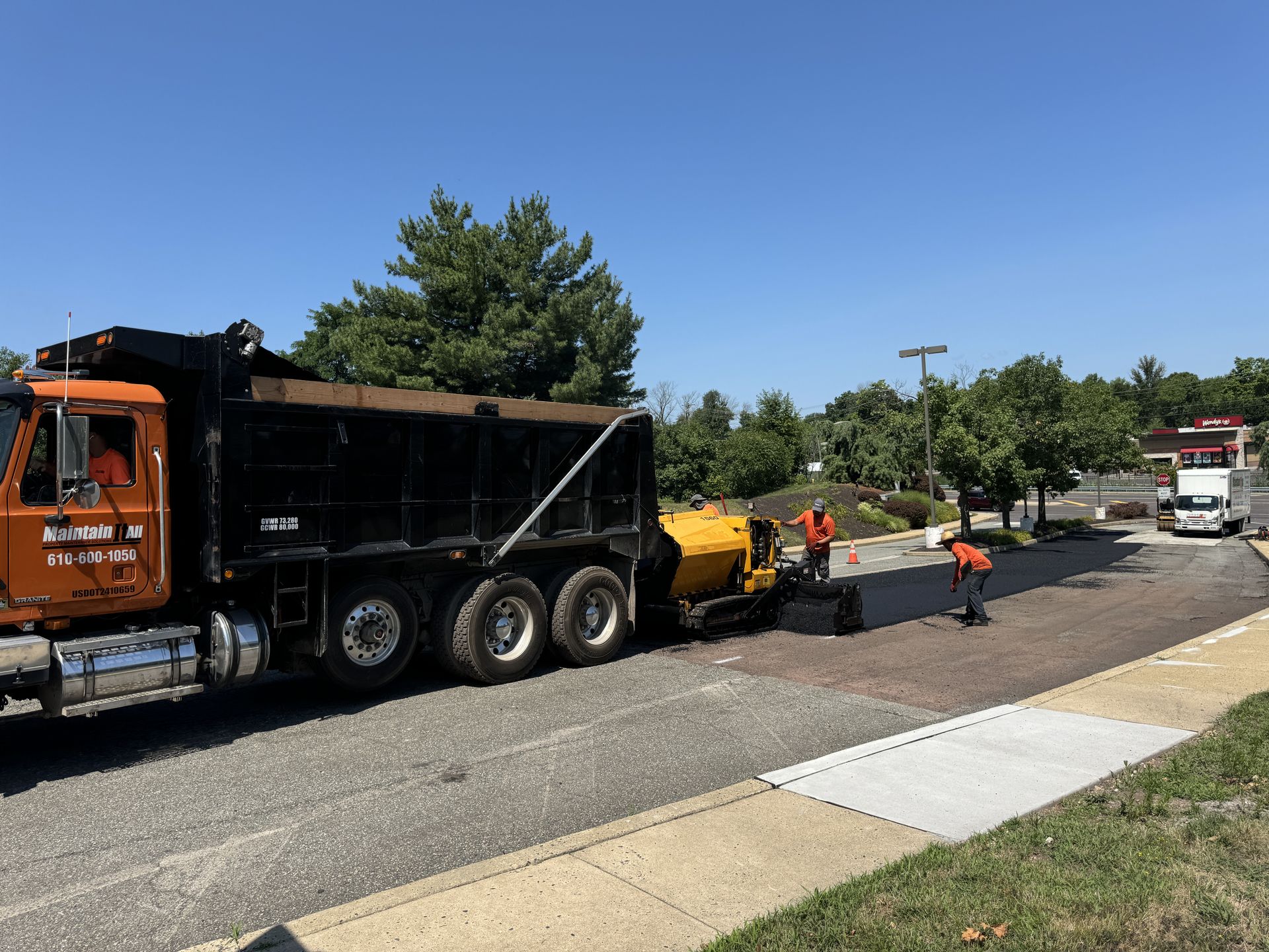 Workers in safety gear use a paving machine and dump truck to resurface an asphalt road on a sunny day.