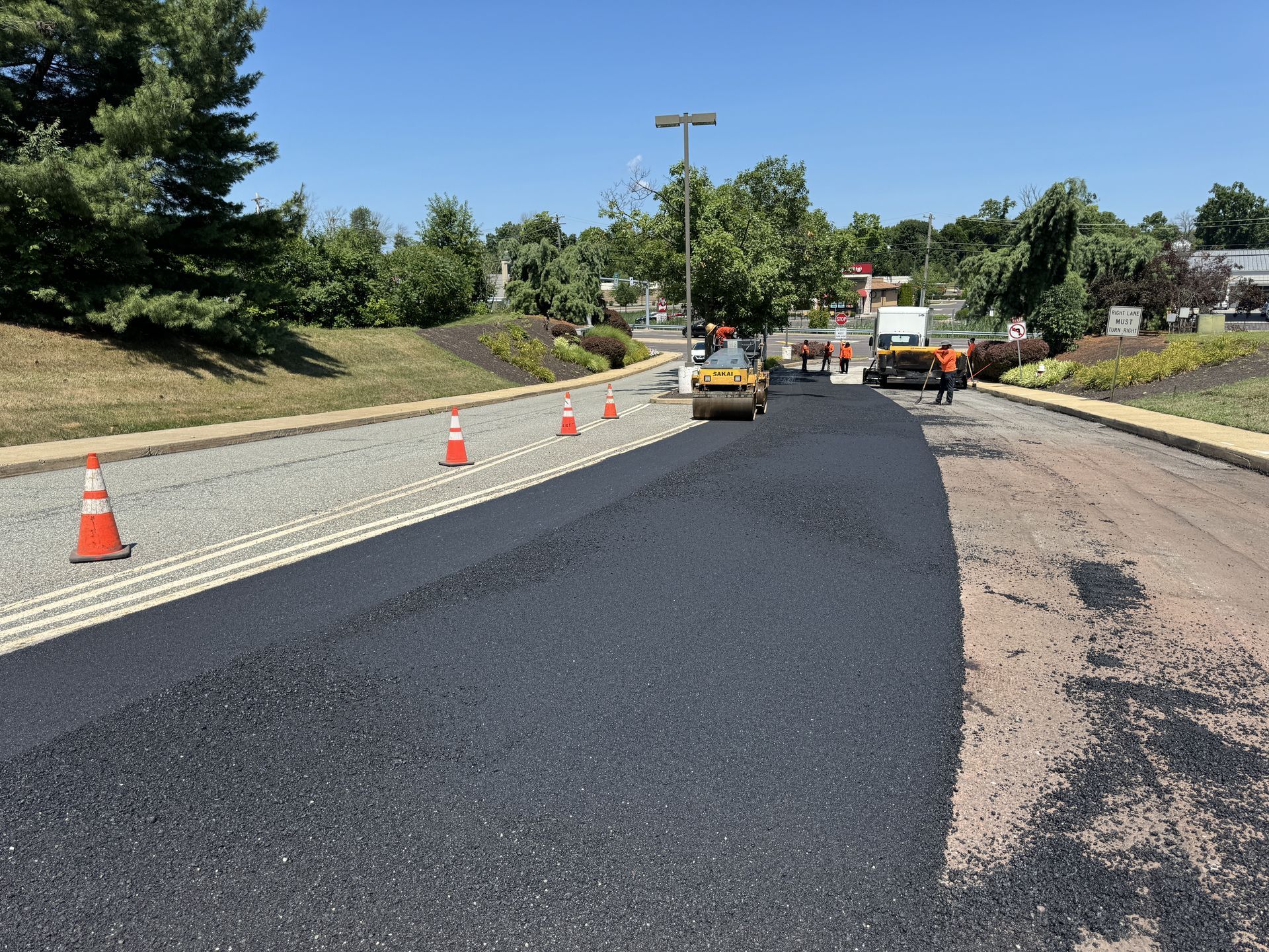 A yellow steamroller paves a road with fresh black asphalt, surrounded by orange traffic cones under a clear blue sky.