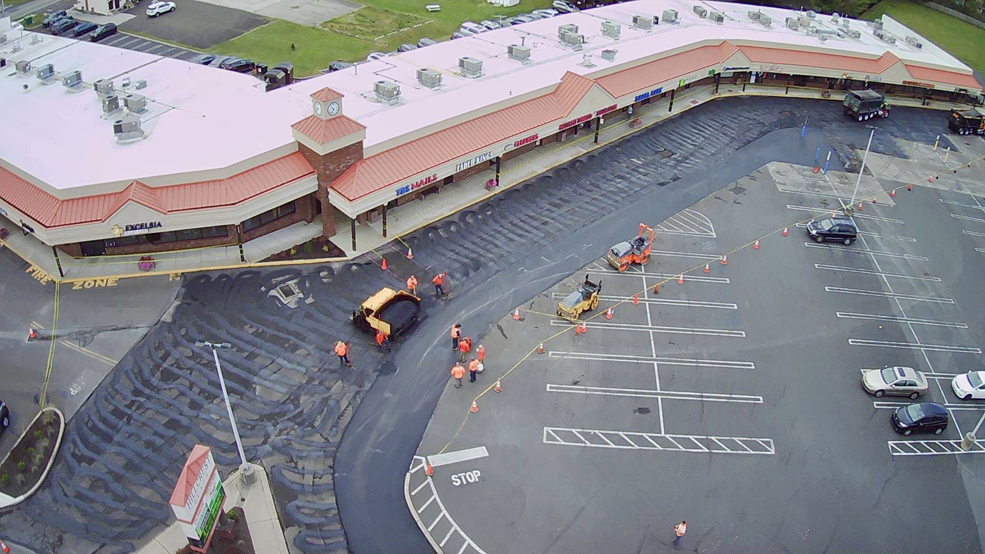 Aerial view of a crew paving an asphalt parking lot in front of a commercial shopping center.