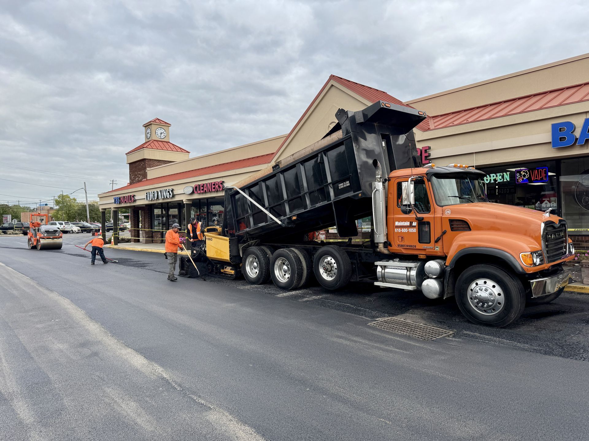 An orange dump truck deposits asphalt onto a parking lot in front of a commercial shopping plaza.