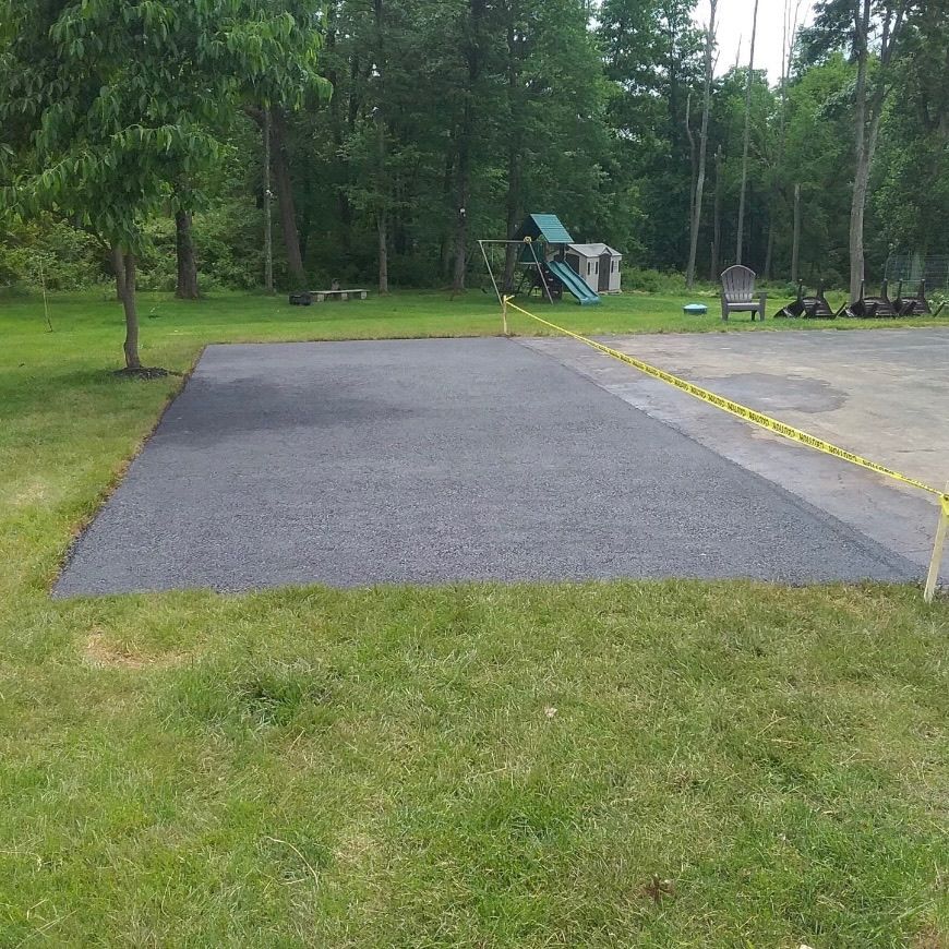 A freshly paved rectangular asphalt driveway patch sits in a grassy yard, bordered by caution tape and older pavement.