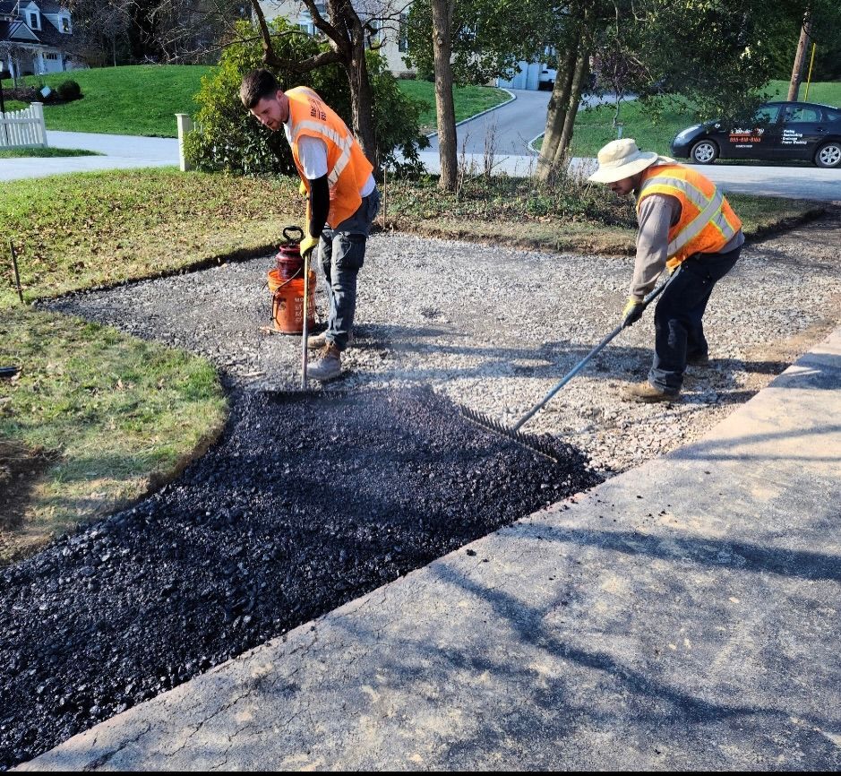 Two workers in orange reflective vests lay new asphalt on a gravel residential driveway.