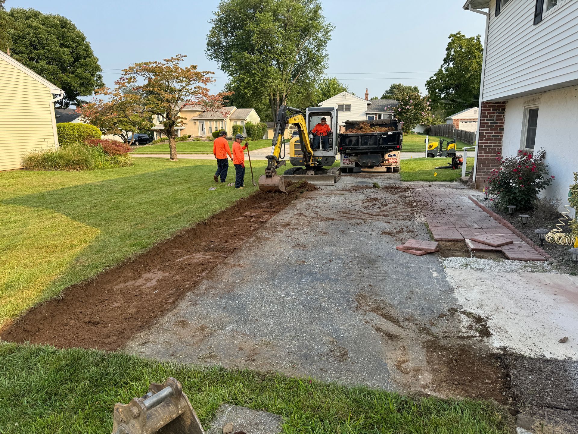 Workers use an excavator to remove paving material along the edge of a driveway next to a house.