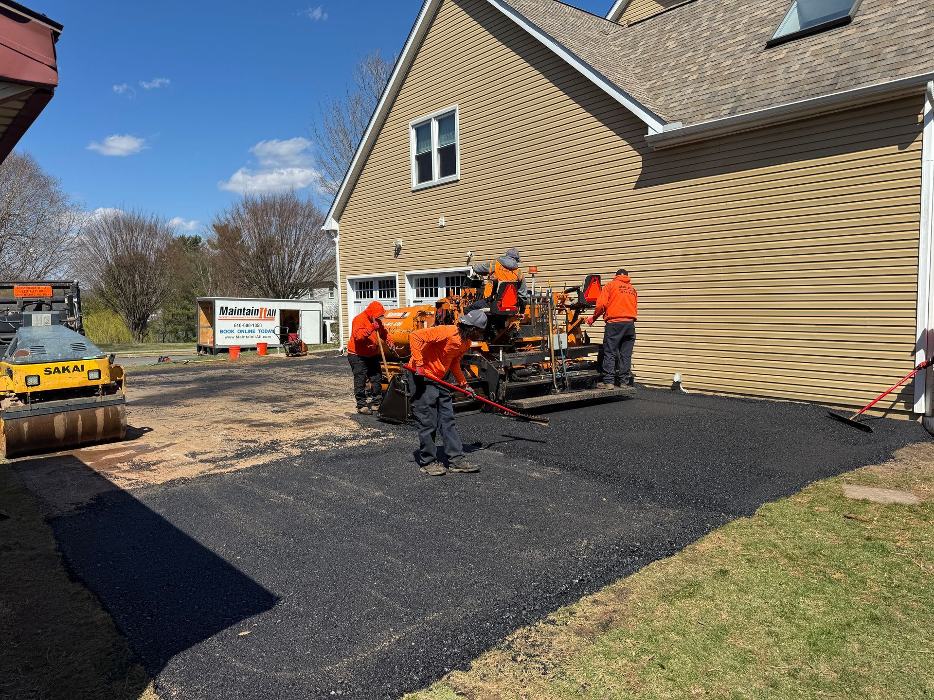 Construction workers in orange safety gear operate heavy machinery to pave a driveway next to a tan house.