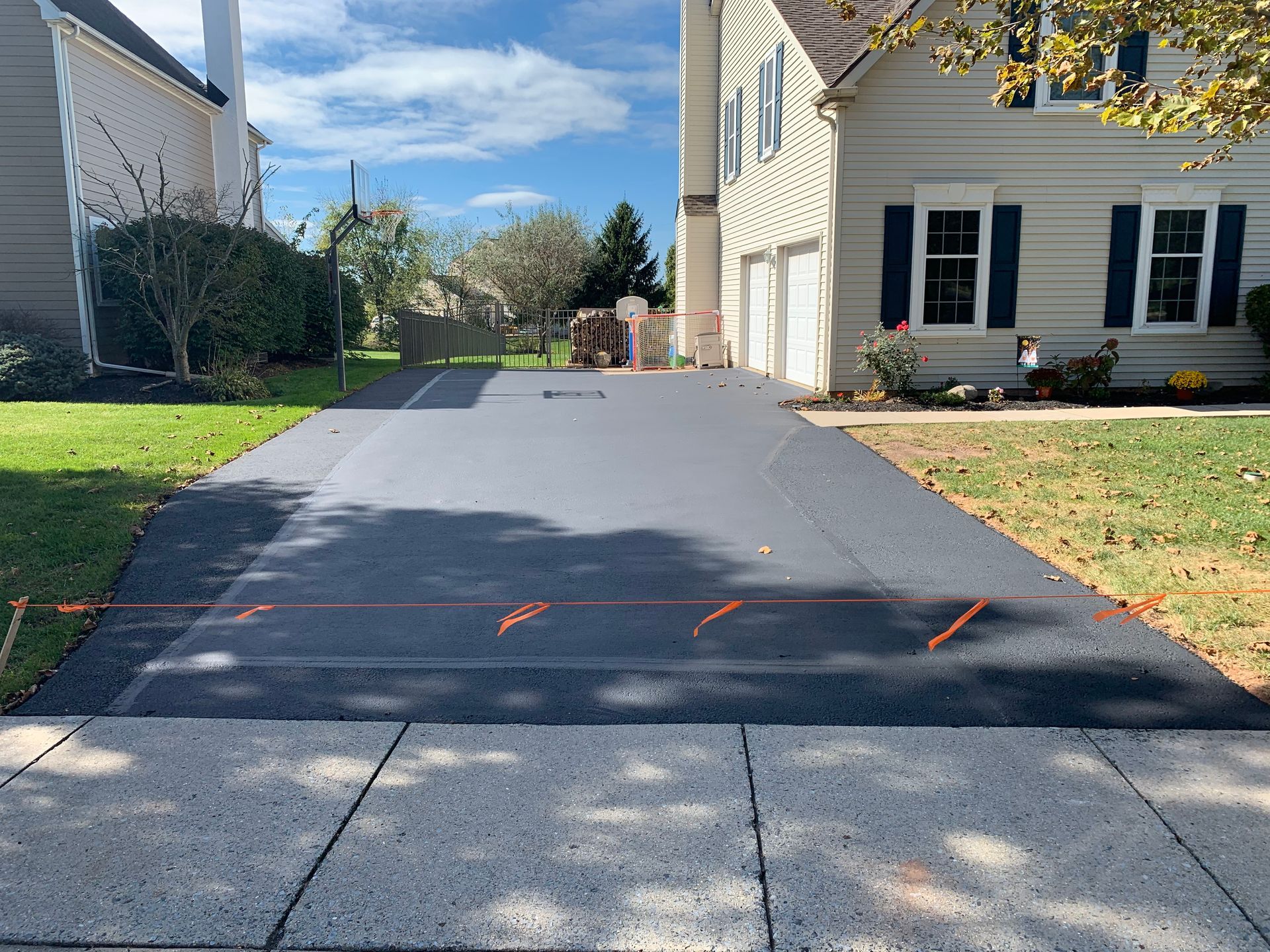 A newly paved driveway leads to a suburban home, with bright orange markings visible on the fresh asphalt.