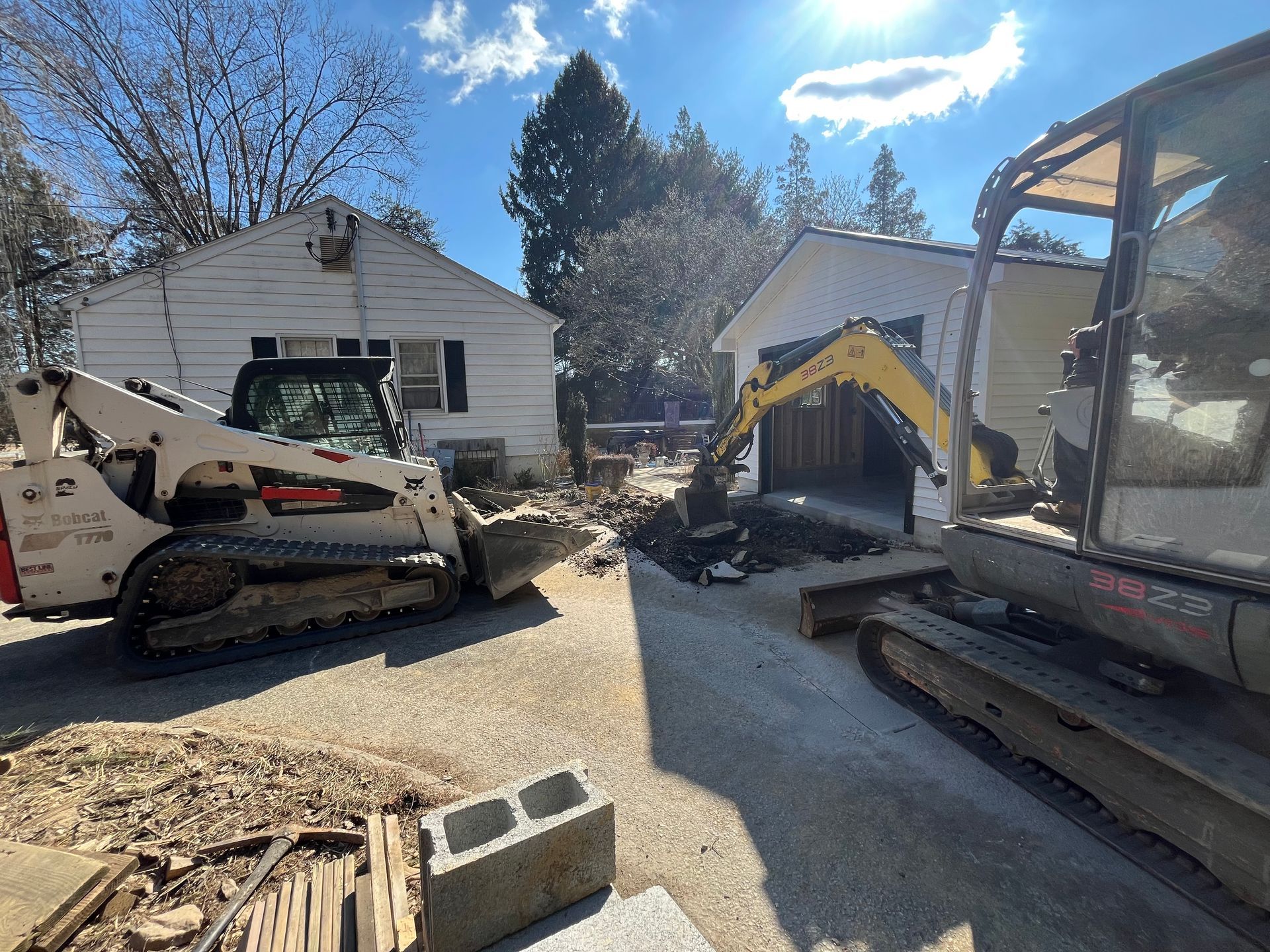 Construction equipment, including a skid steer and excavator, works to clear debris in front of two white buildings.