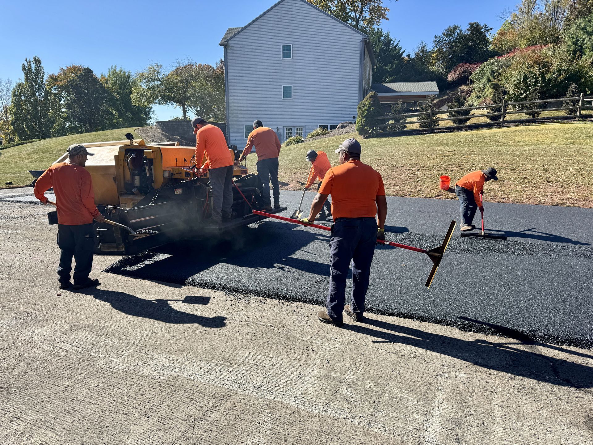 A crew in orange shirts works on paving a residential driveway with fresh asphalt, using a small yellow paver.