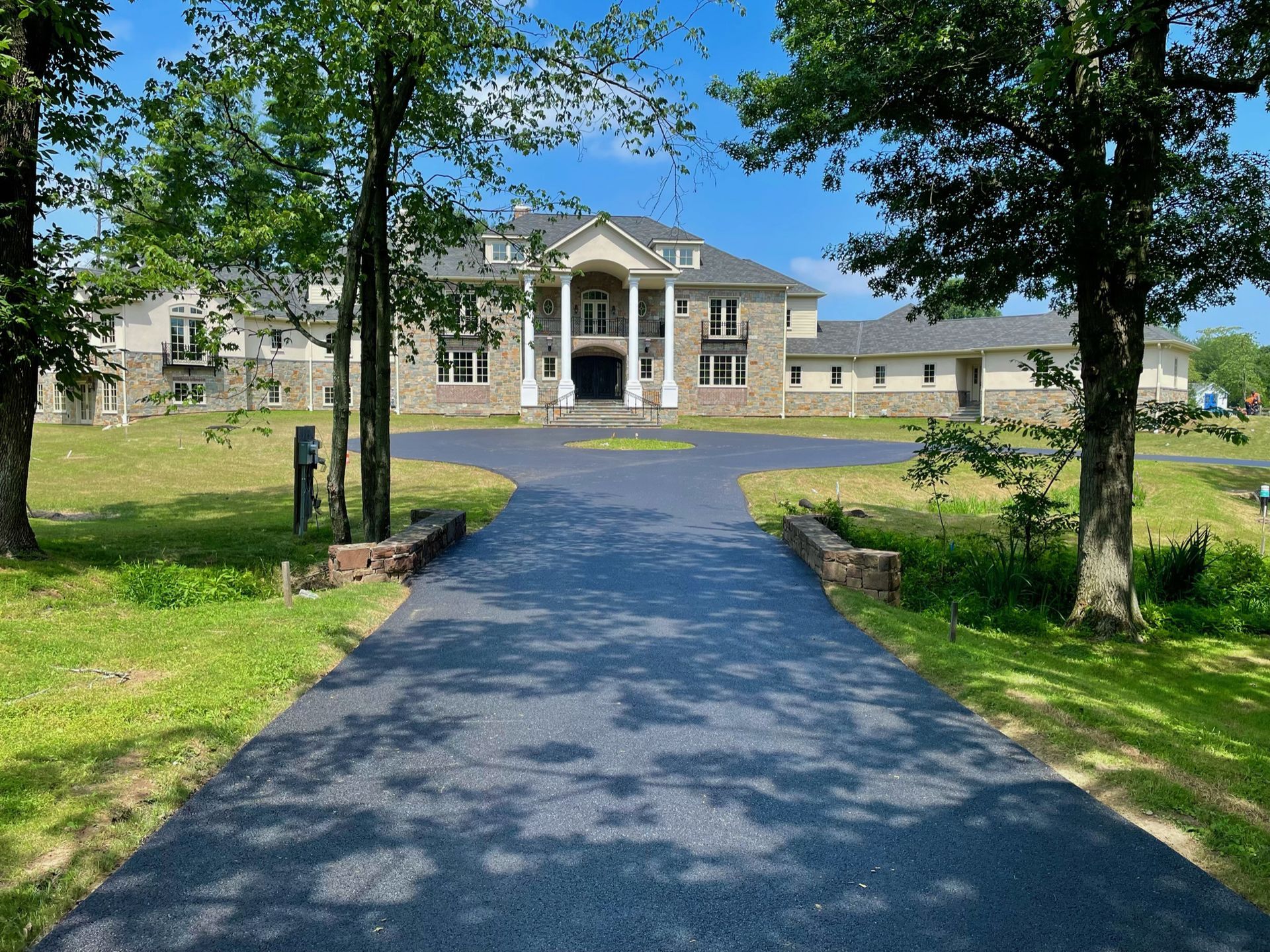 A large, stone-faced two-story mansion with a grand pillared entrance, viewed from the end of a freshly paved driveway.