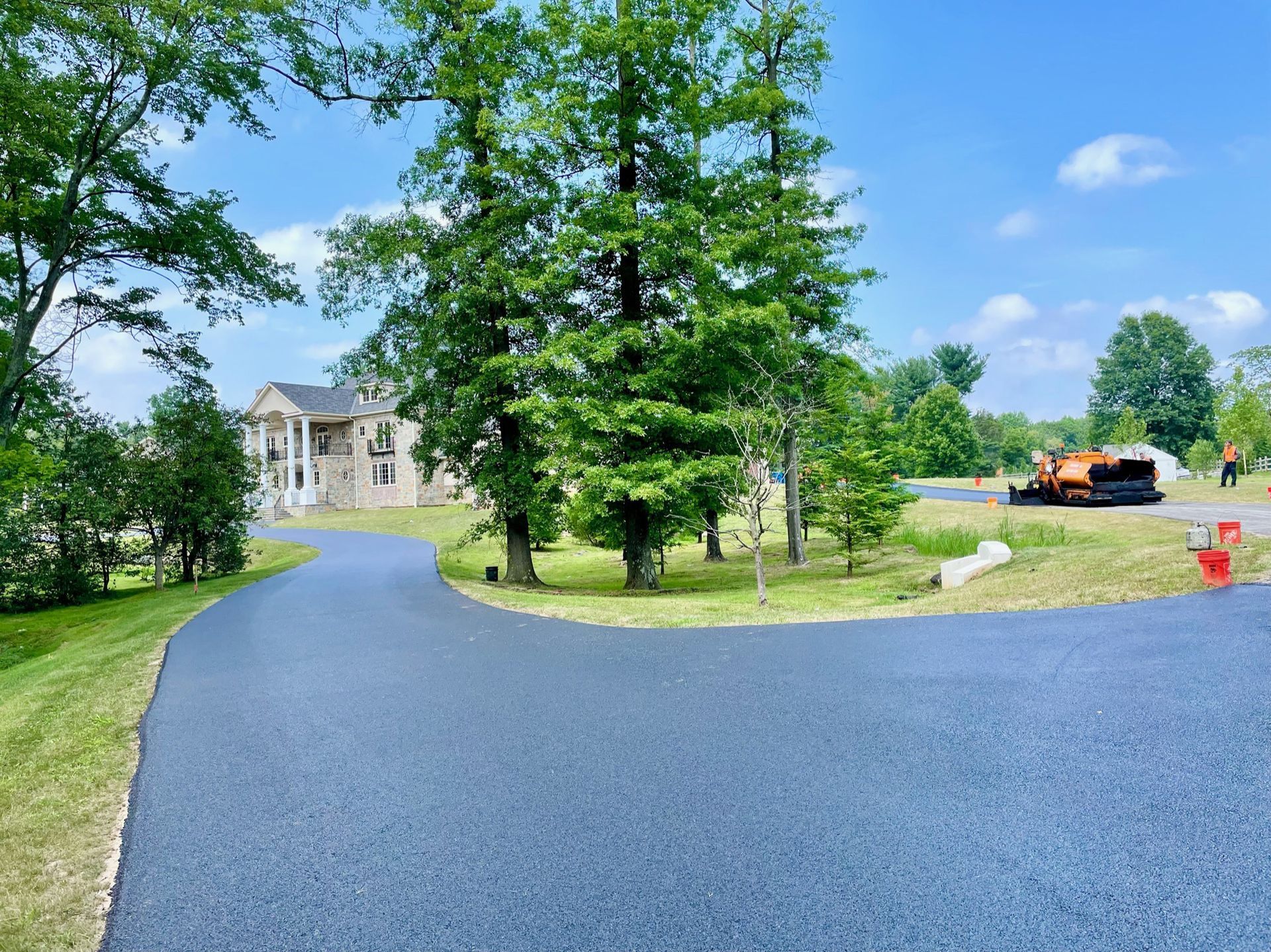 Freshly paved asphalt driveway leading toward a large house surrounded by green trees under a bright blue sky.