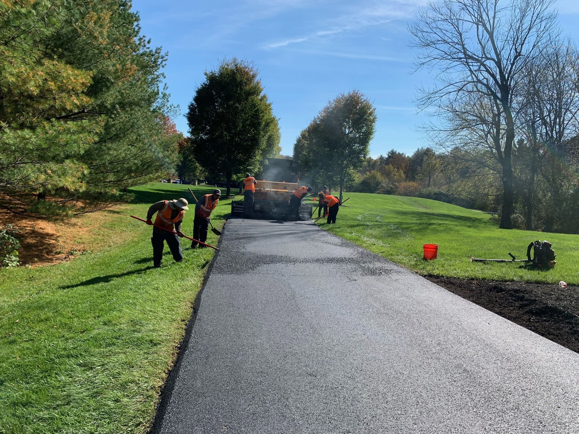 A road construction crew in orange high-visibility vests works on a newly paved asphalt path in a sunny, grassy park.